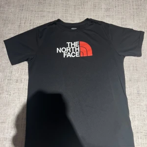 Svart t-shirt från The North Face - Snygg svart t-shirt från The North Face med deras ikoniska logga i vitt och rött på bröstet. Perfekt för en casual look. Tillverkad i ett mjukt och bekvämt material.
