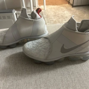 Nike Vapormax i ljusgrått - Säljer ett par stilrena Nike Vapormax i ljusgrått med en unik design utan snörning. Skorna har en synlig luftenhet i sulan för extra komfort och en röd detalj på hälen.