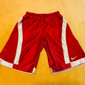 Röda sportshorts från Nike stl S - Snygga röda BB shorts från Nike med vita detaljer och elastisk midja. Perfekta för träning eller bara relaxa i. De har en klassisk design med Nike-loggan på benet. Skönt glansigt material! Storlek XS eller S, märkta 170.