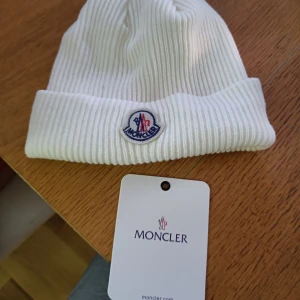 Vit ribbad mössa från Moncler - Snygg vit ribbad mössa från Moncler med broderad logga framtill. Perfekt för kyliga dagar och ger en stilren look. Mössan är i one size och passar de flesta huvudstorlekar.
