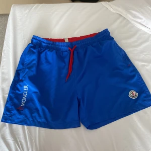Blå badshorts från Moncler - Snygga blå badshorts från Moncler med röd snörning och elastisk midja. Shortsen har Moncler-logga på sidan och en liten broderad detalj. Perfekta för sommarens stranddagar! Vid minsta lilla fundering så hör av er kan handla om vad som helst!