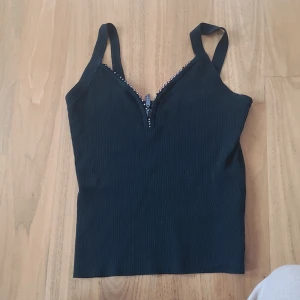 Svart ribbad linnetopp från Beautiful Woman - Snygg svart ribbad linnetopp från Beautiful Woman med dekorativa stenar längs v-ringningen. Några stenar saknas men annars väldigt vacker. 