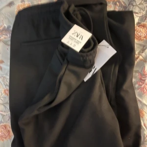 Svarta byxor från Zara - Snygga svarta byxor från Zara med elastisk midja och snörning. Perfekta för en avslappnad stil. Byxorna har en rak passform och är tillverkade i ett mjukt material. Zara tagen är dock borttagen 