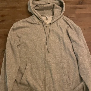 Grå hoodie med dragkedja - Säljer en klassisk grå hoodie med dragkedja och huva. Tröjan har långa ärmar och praktiska fickor framtill. Perfekt för en avslappnad stil.