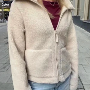 Beige teddyjacka med dragkedja - Mysig beige teddyjacka med dragkedja framtill. Perfekt för kyliga dagar med sin mjuka och varma känsla. Jackan har långa ärmar och två praktiska fickor.