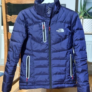 Blå dunjacka från The North Face - Snygg blå dunjacka från The North Face med dragkedja och praktiska fickor. Jackan är tillverkad i nylon och polyester, vilket gör den både vindtät och lätt. Perfekt för kyliga dagar med sin quiltade design och justerbara huva. knappt använd