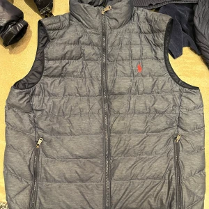 Grå dunväst från Polo Ralph Lauren - Säljer en stilren grå dunväst från Polo Ralph Lauren med dragkedja och två praktiska fickor. Västen har en liten röd logga på bröstet och är perfekt för lager-på-lager under kyliga dagar.