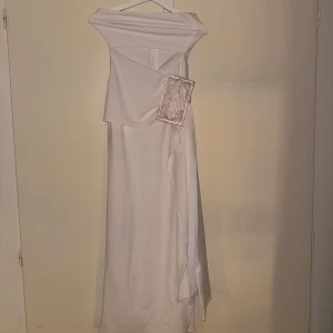 Vit axelbandslös långklänning - Elegant vit långklänning med axelbandslös design och asymmetriska detaljer. Klänningen har en stilren och modern look, perfekt för speciella tillfällen.