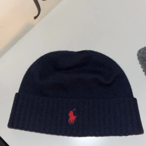 Mörkblå stickad mössa från Ralph Lauren - Snygg mörkblå stickad mössa från Ralph Lauren med röd broderad logga framtill. Perfekt för kyliga dagar och en stilren look.