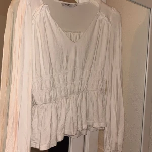 Vit blus från NA-KD - Säljer en stilren vit blus från NA-KD med v-ringning och långa ärmar. Blusen har en bra passform med elastiska detaljer i midjan och volang nedtill. Använd många gånger.