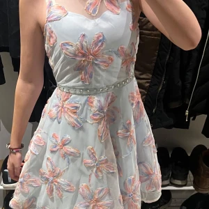 Blommig klänning med pärldetaljer - Charmig ljusblå klänning med stora blommor i rosa och orange. Klänningen har en pärlbeströdd midja och är ärmlös med en söt rosett på axeln. Perfekt för festligheter.