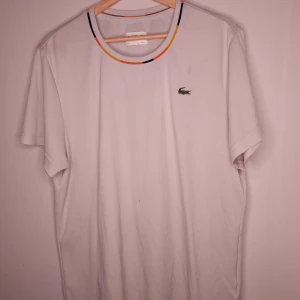 Vit t-shirt från Lacoste Sport - Säljer en vit t-shirt från Lacoste Sport i storlek XL. Den har en färgglad randig krage och det klassiska krokodilmärket på bröstet, i gummi istället för klassiska tyg krokodilen pga att det är "Sport". Materialet är Ultra Dry, perfekt för träning eller varma dagar. T-shirten är i mycket bra skick och redo för nya äventyr!