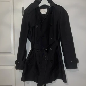 Svart trenchcoat med bälte - Säljer en stilren svart trenchcoat i mycket bra skick. Den har långa ärmar och ett bälte i midjan för en snygg passform. Perfekt för höst och vår! Jackan har knappar framtill och en avtagbar huva. Passar både till vardags och fest.