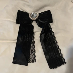 Svart rosett med spets och pärlor - Fin svart stor rosett dekorerad med spets och en glittrig pärla i mitten. Perfekt för att piffa upp en outfit till Rosetten har långa band med spetsdetaljer som ger en elegant touch. Från märket Princess Melody, kan bäras i håret eller sättas fast på en blus i kragen! 