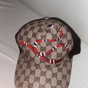 Gucci keps med ormmönster - Säljer en snygg Gucci keps med det klassiska GG-mönstret och en cool röd orm som slingrar sig över framsidan. Kepsen har en svart mesh-baksida för bra ventilation och justerbar rem för perfekt passform. Perfekt för att ge din outfit en lyxig touch!