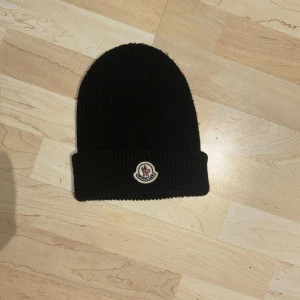 Svart stickad mössa från Moncler - Säljer en snygg svart stickad mössa från Moncler. Den är perfekt för kalla dagar och har en klassisk design med Moncler-loggan framtill. Mössan är i mycket bra skick och passar de flesta tack vare sin flexibla passform. 