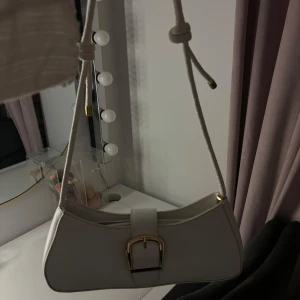 Vit axelväska med gulddetalj - Snygg vit axelväska i syntetmaterial med en elegant guldfärgad spänndetalj på framsidan. Perfekt för både vardag och festliga tillfällen. Väskan har en stilren design och en justerbar axelrem för bästa passform.