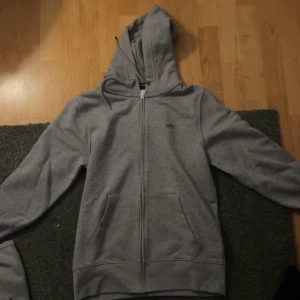 Grå hoodie med dragkedja gant  - Säljer en grå hoodie i bra skick. Den har en dragkedja framtill och en mysig huva. Perfekt för kyliga dagar eller som ett extra lager. Passar både till vardags och avslappnade aktiviteter.