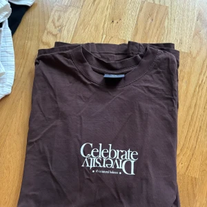 Brun t-shirt med trädmotiv - Säljer en brun t-shirt med texten 'Celebrate Diversity' och ett coolt trädmotiv på baksidan. Perfekt för dig som gillar naturen och vill ha en avslappnad stil. T-shirten är i bomull och har en normal passform. Passar perfekt till vardags eller en chill dag ute. Står som storlek S men är mer som M. 