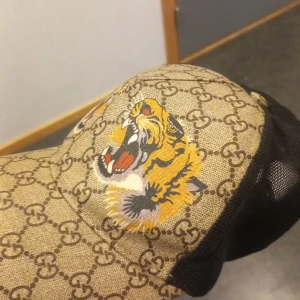 Gucci keps med tigerbroderi - Säljer en snygg Gucci keps med ett coolt tigerbroderi på sidan. Kepsen har det klassiska Gucci-mönstret i beige och brunt, och ett justerbart band i grönt och rött baktill. Perfekt för att ge din outfit en lyxig touch!