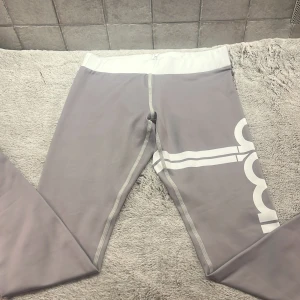 Grå leggings från Aim'n - Säljer ett par grå leggings från Aim'n i storlek XS. De är tillverkade i 73% polyester och 27% spandex, vilket gör dem superstretchiga och bekväma. Perfekta för träning eller en chill dag hemma. De har en hög midja och en snygg vit detalj på benet. Dom är i ny skick
