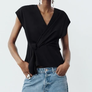 Svart omlottopp från Zara - Snygg svart omlottopp från Zara i ett elegant och stilrent utförande. Toppen har en v-ringad design och är ärmlös, vilket gör den perfekt för både vardag och fest. Materialet känns mjukt och bekvämt mot huden.