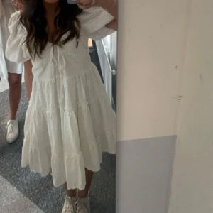 Vit klänning från Chiquelle - Superfin vit klänning från Chiquelle i broderad bomull. Den har korta puffärmar och en söt knytning framtill. Perfekt för sommarens alla fester eller en mysig dag på stan. Klänningen har en luftig passform och volanger som ger en romantisk känsla. 🕊️