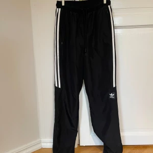 Svarta trackpants träningsbyxor från Adidas - Säljer ett par svarta träningsbyxor från Adidas i mycket bra skick. De har de klassiska vita ränderna längs sidorna och en bekväm resårmidja med snörning. Perfekta för träning eller en avslappnad dag. Adidas-loggan syns på ena benet. Passar till både höst och vår.