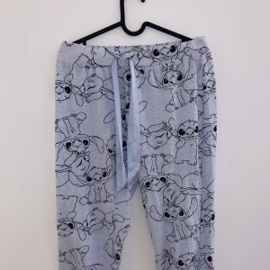 Grå mjukisbyxor med Stitch-mönster - Supermysiga grå mjukisbyxor med ett gulligt Stitch-mönster över hela tyget. Perfekta för en avslappnad dag hemma eller som pyjamasbyxor. De har en bekväm resår i midjan med snörning för bästa passform. Materialet känns mjukt och skönt mot huden.