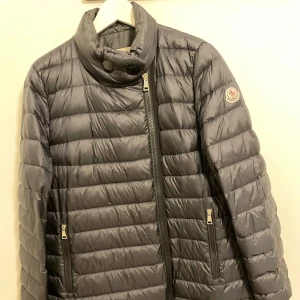 Navy dunjacka från Moncler - Säljer en snygg grå dunjacka från Moncler i mycket bra skick. Jackan har en quiltad design med dragkedja och knappar vid kragen. Perfekt för höst och vinter, håller dig varm och stilren. 