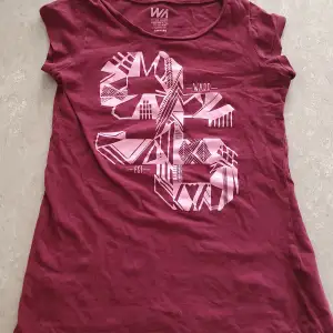 Säljer en cool vinröd t-shirt med ett stort grafiskt tryck i rosa på framsidan. T-shirten är kortärmad och gjord i mjukt bomullsmaterial, perfekt för vardagsbruk. Passar bra till jeans eller shorts för en avslappnad stil.