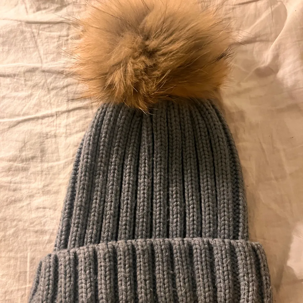 Superfin grå stickad mössa med en stor fluffig pälsboll på toppen. Perfekt för kalla vinterdagar när du vill hålla dig varm och se stilren ut. Den ribbade designen ger en skön passform och extra värme. Ett måste för vinterns garderob!. Asusteet.