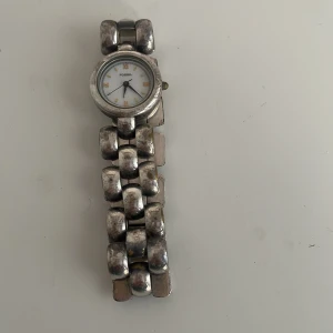 Silverfärgad klocka från Fossil - Säljer en stilren klocka från Fossil i silverfärg. Den har en unik länkdesign och en vit urtavla med guldiga detaljer. Perfekt för både vardag och fest. Klockan är i acceptabelt skick med lite slitage på länken. Passa på att köpa denna tidlösa accessoar!