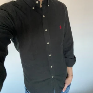 Ralph Lauren skjorta - Säljer en stilren svart skjorta från Ralph Lauren i slim fit. Den är perfekt för både vardag och fest. Den röda loggan på bröstet ger en snygg kontrast. Passar bra till jeans eller chinos för en klassisk look. Hör gärna av er vid funderingar eller frågor!