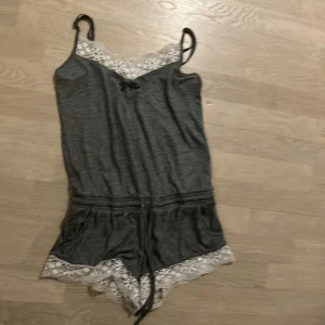 Grå jumpsuit med spetsdetaljer - Säljer en superfin grå jumpsuit med vita spetsdetaljer vid halsringningen och bensluten. Den har smala axelband och en knytning i midjan för en bekväm passform. Perfekt för en avslappnad dag hemma eller sommarens varma kvällar.