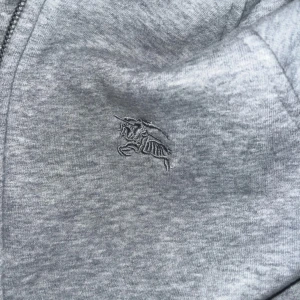 Grå huvtröja med dragkedja - Säljer en snygg grå huvtröja från Burberry i storlek XS. Den har en dragkedja framtill och en klassisk rutig mönster i huvan. Materialet är en blandning av bomull och polyester, vilket gör den både mjuk och hållbar. Perfekt för höst och vår! Skicken är 10/10.