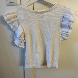 Vit ribbad topp från Zara - Säljer en superfin vit ribbad topp från Zara. Den har korta ärmar med volangdetaljer som ger en söt touch. Perfekt för både vardag och fest! 🕊️