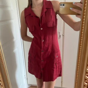 Vinröd ärmlös skjortklänning - Snygg vinröd skjortklänning i linnematerial, ärmlös med röda knappar och fickor. Köpt second hand men aldrig använd. Mini-längd. En knapp saknas högst upp  (se sista bild) men påverkar i min upplevelse inte passformen. Hör gärna av dig för mer info!