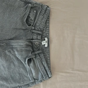 Grå jeansbyxor - Säljer ett par grå jeansbyxor i bra skick. De har en klassisk femficksdesign med två bakfickor och tre framfickor, inklusive en myntficka. Byxorna har en knapp och dragkedja framtill. Perfekta för vardagsbruk och kan matchas med nästan vad som helst. Passar både till skola och fritid.