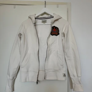 Vit hoodie från Polo Jeans Company - Säljer en snygg vit hoodie från Polo Jeans Company (Ralph Lauren). Den har en dragkedja framtill och en cool broderad logga med siffran 67 på bröstet. Perfekt för en avslappnad stil och passar till det mesta. Den är i bra skick (lite nopprig) och redo för nya äventyr! Lite liten i storleken.