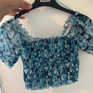 Blommig topp från KappAhl - Säljer en superfin blommig topp från KappAhl. Den är blå och svart med ett smockat mönster och korta puffärmar. Toppen är croppad och har en söt volangkant. Perfekt för sommardagar eller en festlig kväll!