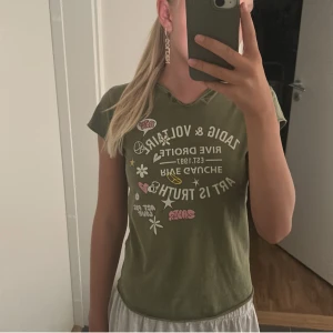 Grön t-shirt från Zadig & Voltaire - Säljer en cool grön t-shirt från Zadig & Voltaire. Den har ett tryck på framsidan med texten 'Art is Truth' och olika symboler som blommor, hjärtan och peace-tecken. T-shirten är i bomull och har korta ärmar. Perfekt för en avslappnad och trendig look!