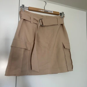Beige kjol från Gina Tricot - Säljer en superfin beige kjol från Gina Tricot i storlek 36. Kjolen har en stilren design med ett bälte i midjan och praktiska fickor på sidorna. Perfekt för både vardag och fest!