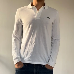 Vit långärmad pikétröja från Lacoste - Säljer en stilren och klassisk vit långärmad pikétröja från Lacoste. Tröjan har den ikoniska krokodilloggan på bröstet och är perfekt för både vardag och lite finare tillfällen. Den är i bra skick och passar perfekt till jeans eller chinos. Skön och bekväm att ha på sig! 