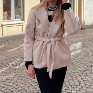 Missäy kappa - Säljer en snygg beige kappa med bälte i midjan. Den har en klassisk design med en bred krage och är perfekt för både höst och vår. Jackan är långärmad och har en normal passform. Perfekt för att hålla sig varm och stilren på samma gång!