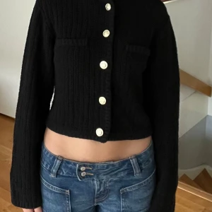 Svart stickad jacka från H&M - Säljer en snygg svart stickad jacka från H&M. Den har en croppad passform och är långärmad. Jackan har fyra knappar framtill och två dekorativa fickor på bröstet. Perfekt för höst och vår!