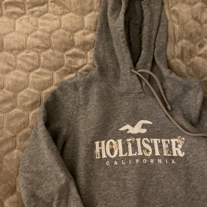 Grå hoodie från Hollister - Säljer en supermysig grå hoodie från Hollister med en snygg broderad logga och blommönster. Perfekt för kyliga dagar och har en skön passform med långa ärmar och en praktisk huva. Perfekt för både vardag och chill.