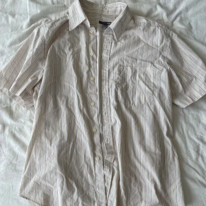 Randig kortärmad skjorta från H&M - Säljer en snygg och bekväm randig kortärmad skjorta från H&M. Skjortan är i storlek L och har ett klassiskt mönster med tunna ränder i beige, svart och orange. Den är tillverkad i ett lätt och luftigt material, perfekt för varma dagar. Skjortan har knappar hela vägen ner och en bröstficka på vänster sida. Perfekt för både vardag och fest! Frågor? Skriv!