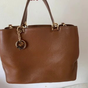 Brun skinnväska från Michael Kors - Säljer en snygg brun skinnväska från Michael Kors. Den har både handtag och en axelrem, vilket gör den superpraktisk. Väskan har en fin gulddetalj med MK-loggan och en dragkedja upptill. Insidan är rymlig med flera fack för att organisera dina grejer. Perfekt för både vardag och fest! 2st band ingår långt och kort + dustbag om det önskas!