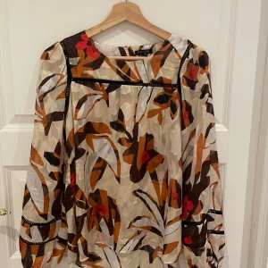 Mönstrad blus med långa ärmar - Säljer en superfin blus med ett färgglatt mönster i beige, brun, orange och röd. Blusen har långa ärmar och en rund halsringning. Perfekt för höst och vår, och den har en bekväm passform som är lite loose. Den är gjord i ett lätt och luftigt material, vilket gör den perfekt för både vardag och fest.